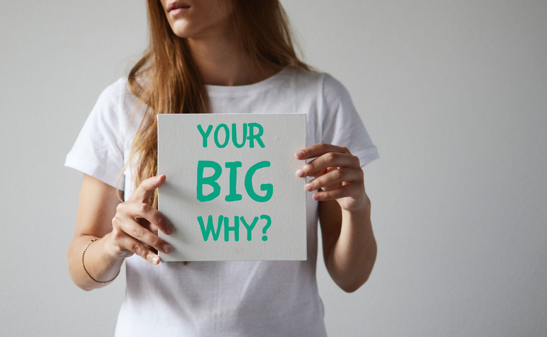 A woman wearing a white t-shirt holding a card that says 'Your Big Why?'