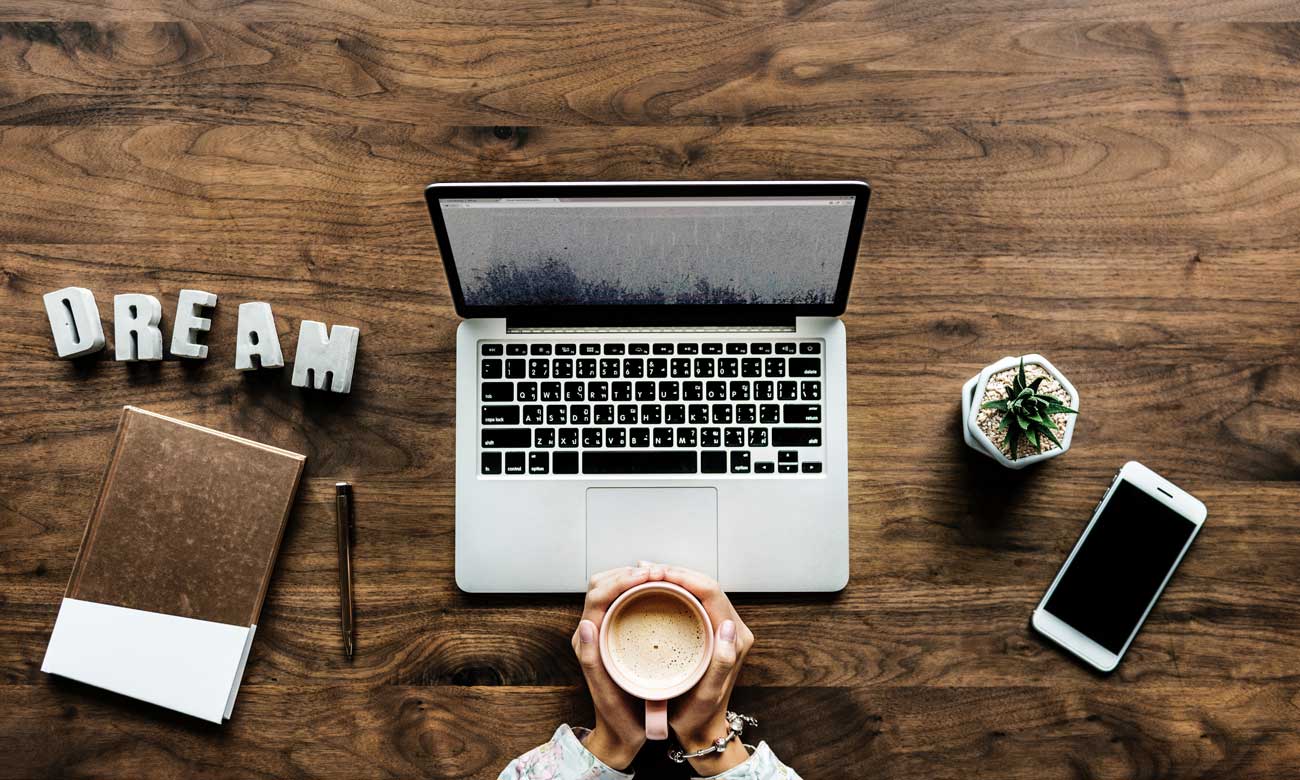 Top-down view of a laptop on a table, a man holding a coffee with two hands, along with a phone and notes.