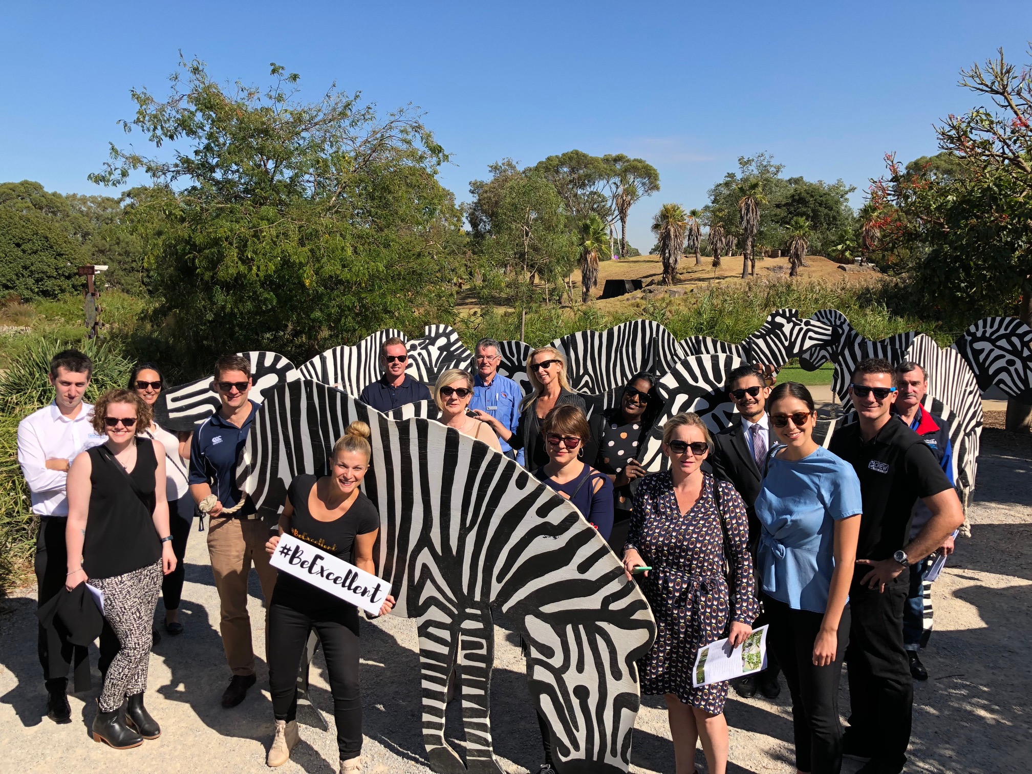 Despina Karatzias holding a #BeExcellent cardboard sign with a group of people in an outdoor field, alongside cut-out images of zebras.