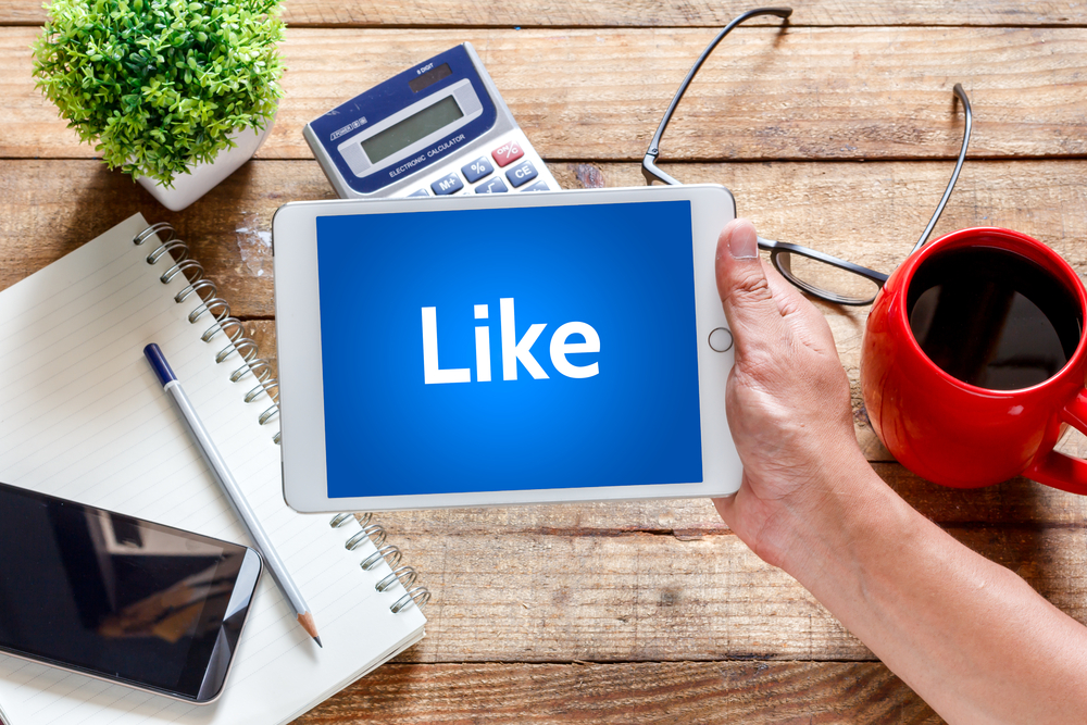 A man holding an iPad displaying the Facebook 'Like' icon, placed on a table with a notebook, coffee cup, and eyeglasses nearby.
