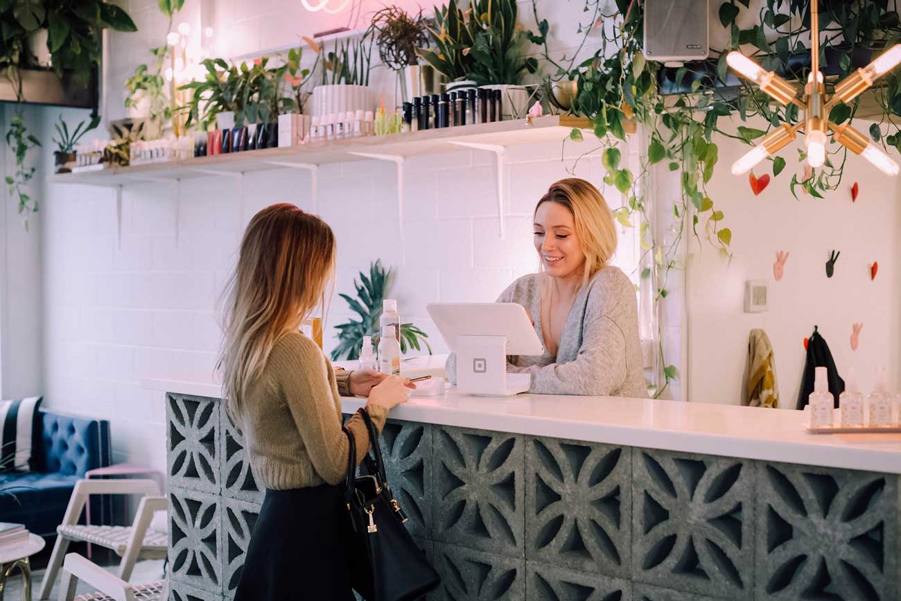 Two women interacting at a stylish reception counter surrounded by greenery and soft lighting.