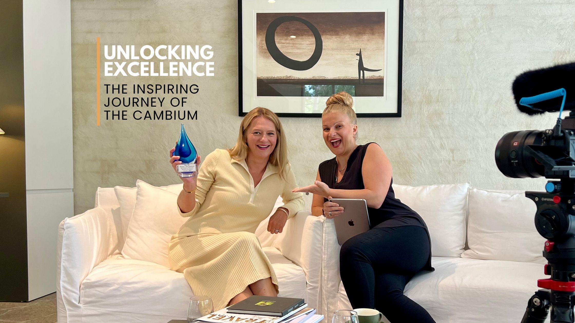 Two women sitting on a white couch, one holding an award and the other holding a tablet, with a camera setup and text overlay: Unlocking Excellence - The Inspiring Journey of The Cambium.
