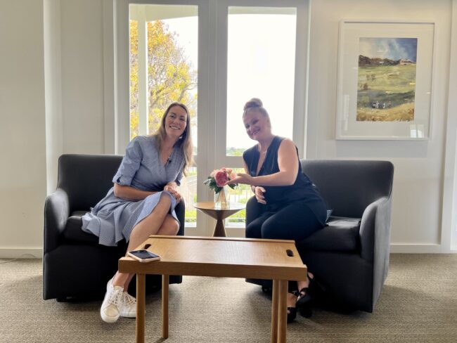 Friendly Conversation in a Bright and Relaxing Setting Two women sitting on couches in a well-lit room, smiling and sharing a moment over a small table with a flower vase.