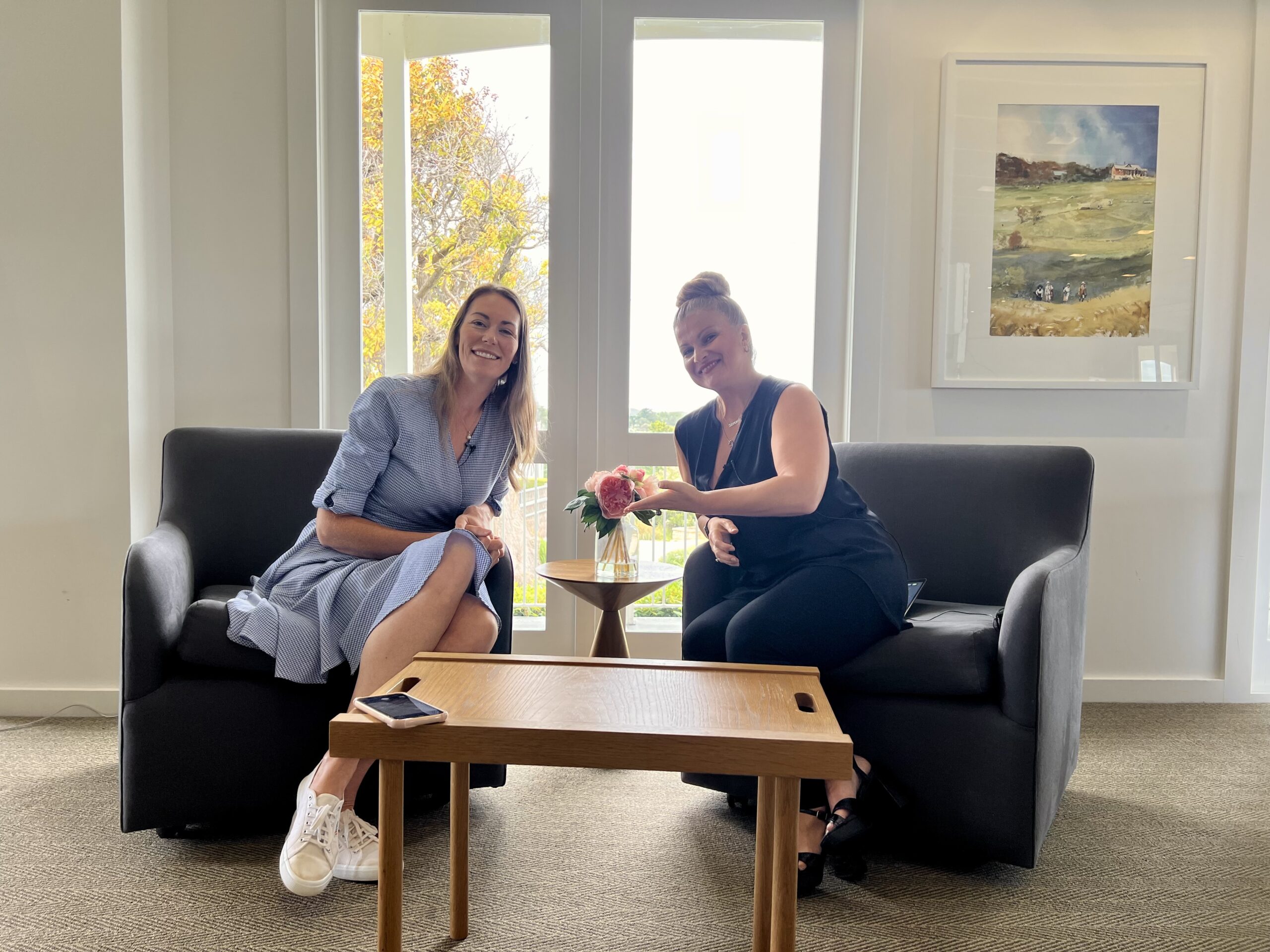 Two women sitting on couches in a well-lit room, smiling and sharing a moment over a small table with a flower vase.
