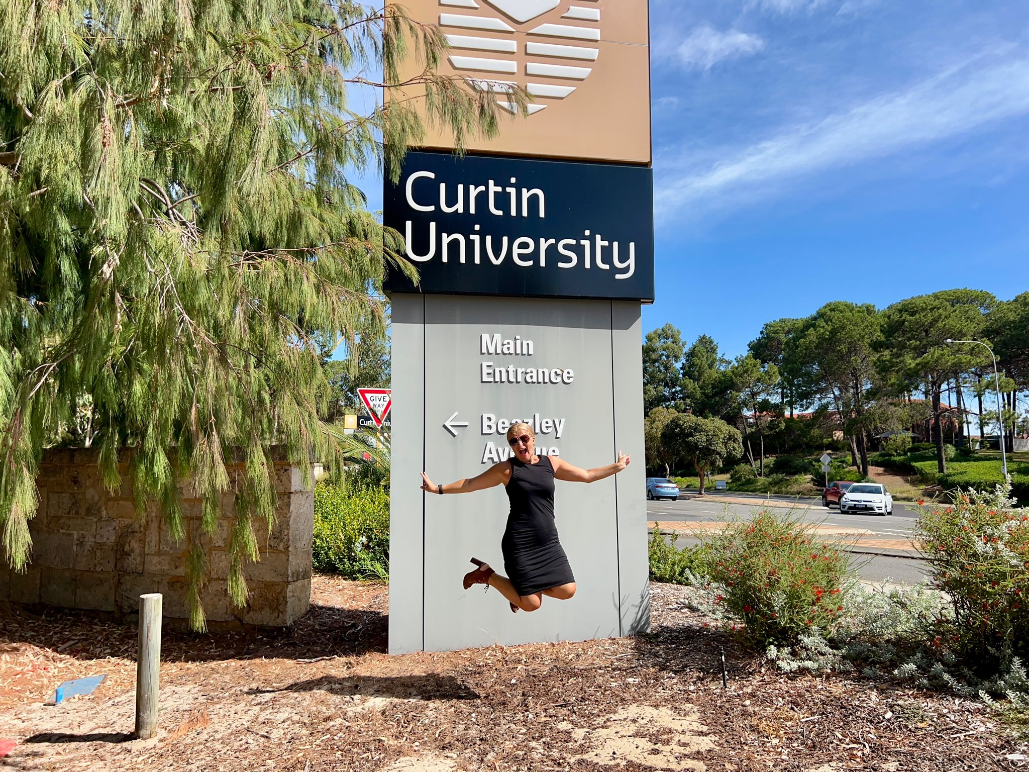 Smiling woman jumping in front of the Curtin University sign with trees and a blue sky in the background.