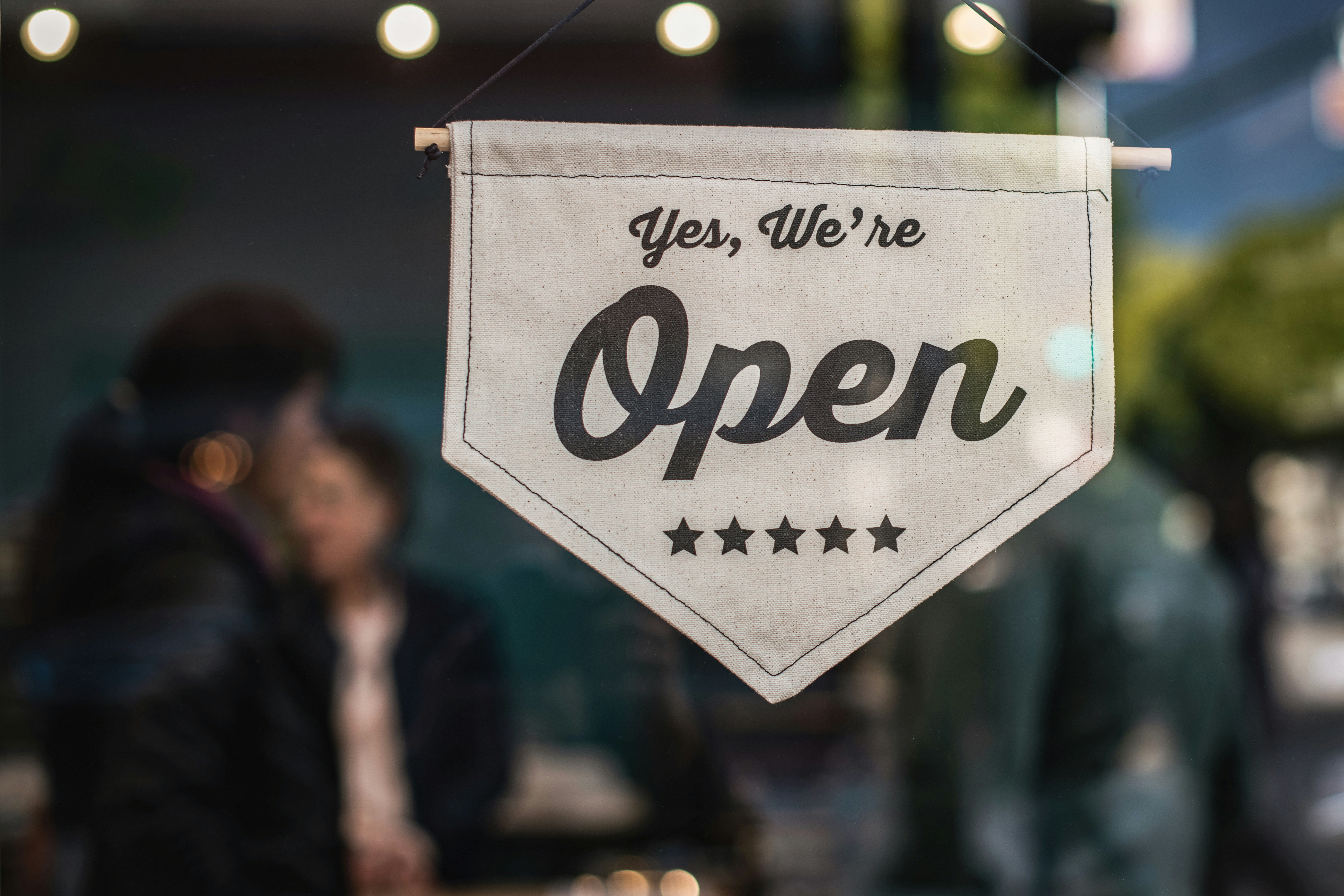 A shop window sign reading “Yes, We’re Open” hanging in front of a blurred background with people inside a small business.