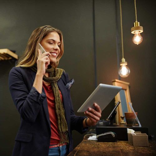 Busy tourism business owner on the phone while checking a tablet at her shop counter.