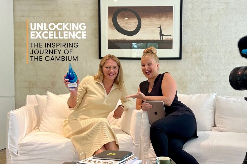 Two women sitting on a white couch, one holding an award and the other holding a tablet, with a camera setup and text overlay: Unlocking Excellence - The Inspiring Journey of The Cambium.