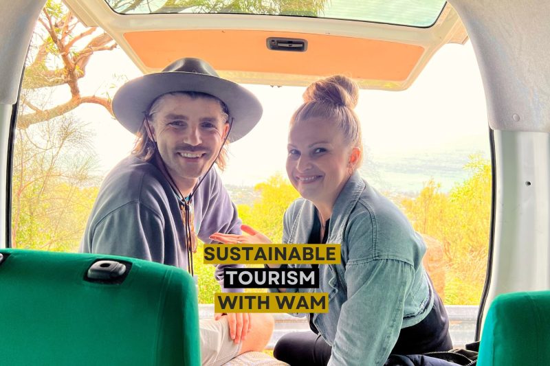 Man in a hat and woman smiling inside a van with text overlay: Sustainable Tourism with WAM, overlooking a scenic landscape.