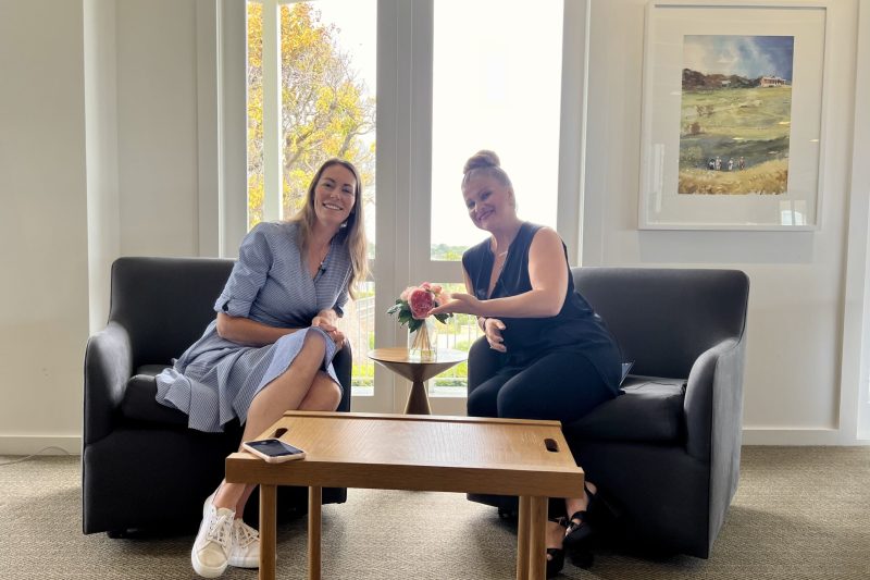 Two women sitting on couches in a well-lit room, smiling and sharing a moment over a small table with a flower vase.