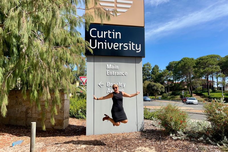 Smiling woman jumping in front of the Curtin University sign with trees and a blue sky in the background.